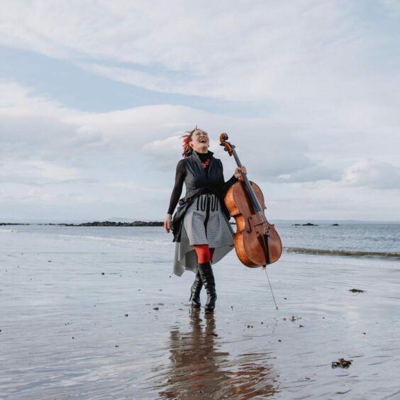 Cellist Su-a Lee is pictured on a beach in Fife. She is holding her cello and her head is thrown back in laughter.