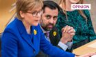 Humza Yousaf seated next to Nicola Sturgeon, who is standing at her desk addressing the Scottish Parliament.