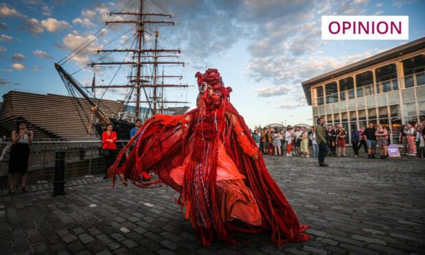 Person draped in red material performing for a crowd of people beside RRS Discovery and the V&A Dundee museum at Dundee waterfront as part of the Art Night event.