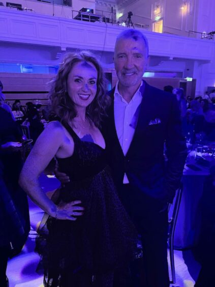 Martel Maxwell in party dress with former footballer Graeme Souness. The pair are posing for the camera in a busy function room at the Pride of Scotland awards, surrounded by tables and fellow guests.