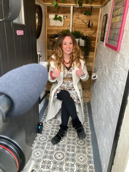 Martel Maxwell sitting on a toilet giving the thumbs up sign while she is filmed for TV's Homes Under The Hammer.