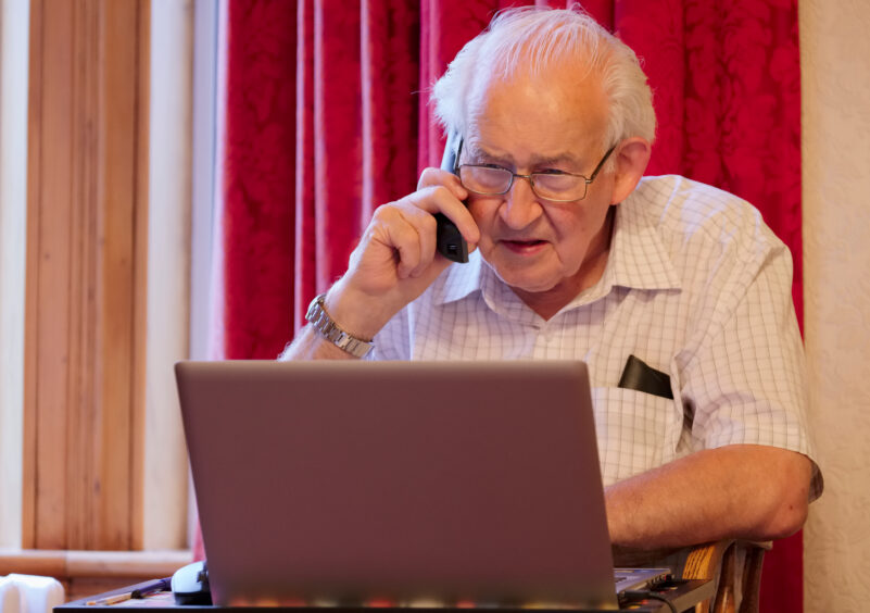 A photo of an elderly man on the phone at his laptop.