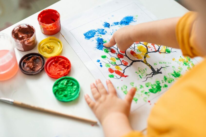 Child finger painting a drawing of a tree, photographed from above, with open paint pots by their side.