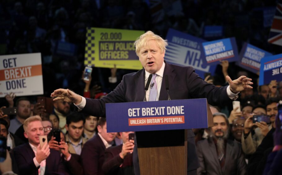 Boris Johnson delivering a speech from behind a lectern with the slogan 'Get Brexit done', surrounded by a large crowd of supporters holding placards and filming on their mobile phones.