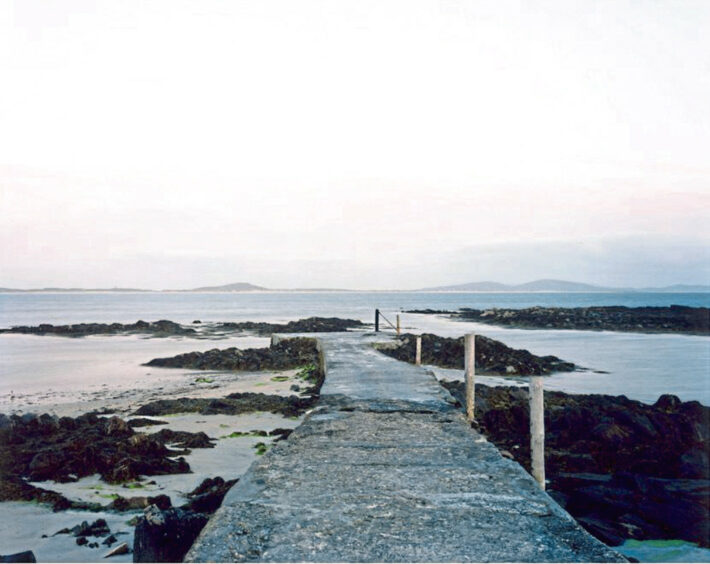 The image shows Harry Cory Wright's photograph of Pabby in Scotland for the Space to Breathe Exhibition. There is a pier jutting out to sea surrounded by rocks and seaweed.