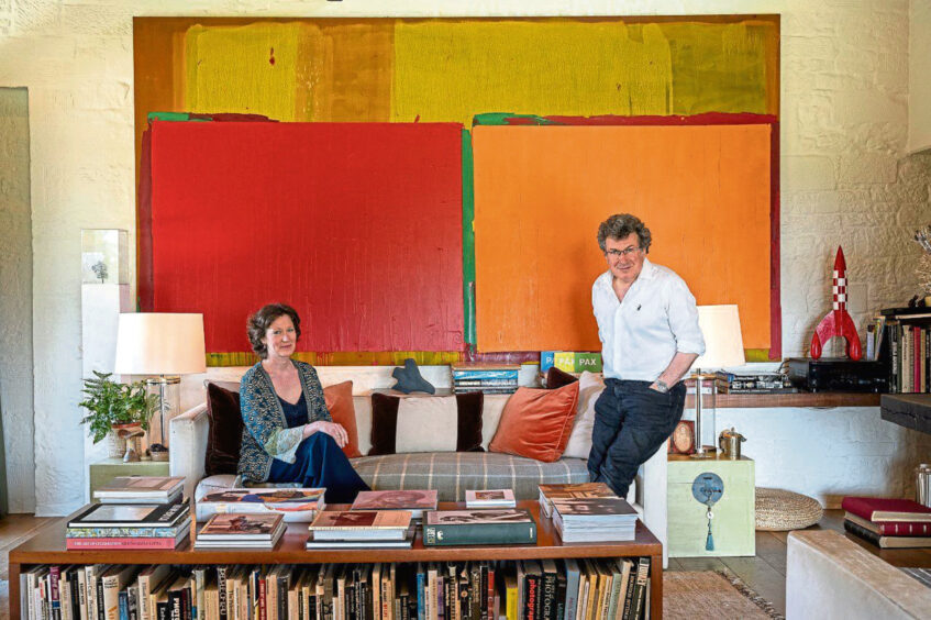 Sophie Camu is pictured sitting with her husband, the photographer Alexander Lindsay standing close by. They are surrounded by books and artworks.
