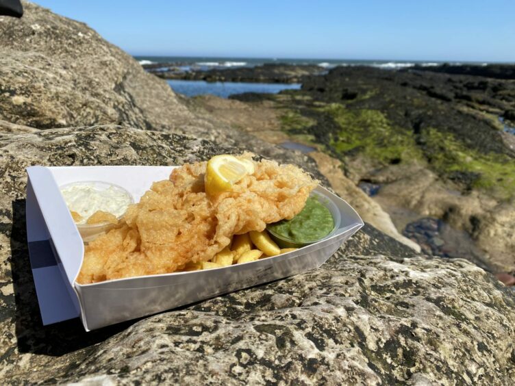 a box of fish and chips from Cromars against the backdrop of the coastline in Fife, UK. 
