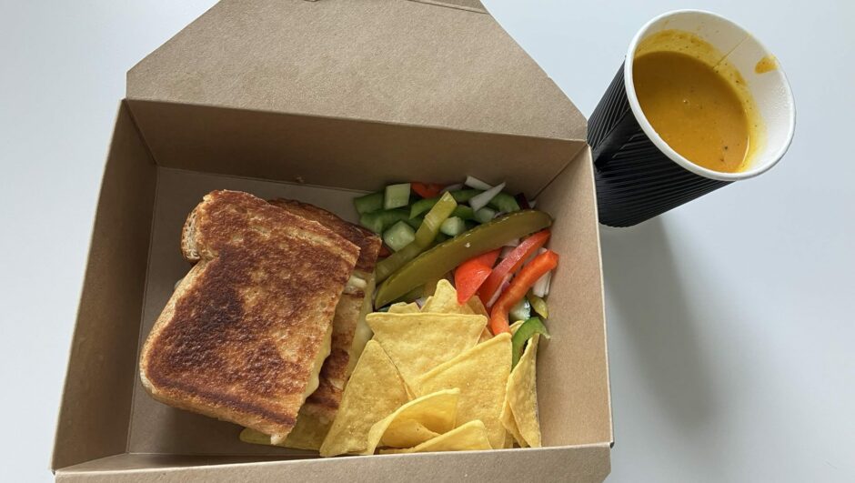 A takeaway box with a cheese toastie, tortilla chips and salad, with a cup of soup on the side.