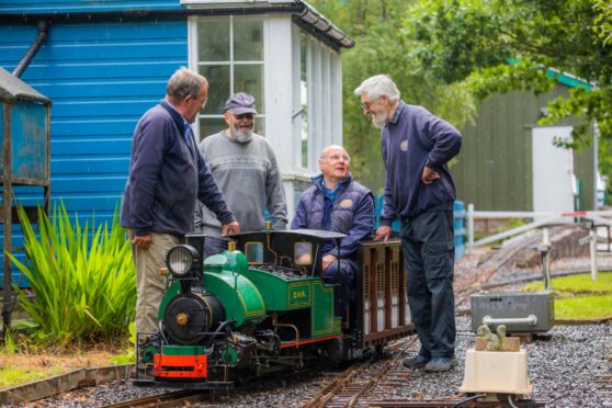 Three men standing around a miniature train with a fourth man in the driver's seat.