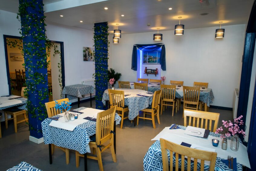A Greek taverna with white walls, blue columns, white and blue tablecloths and wooden chairs.