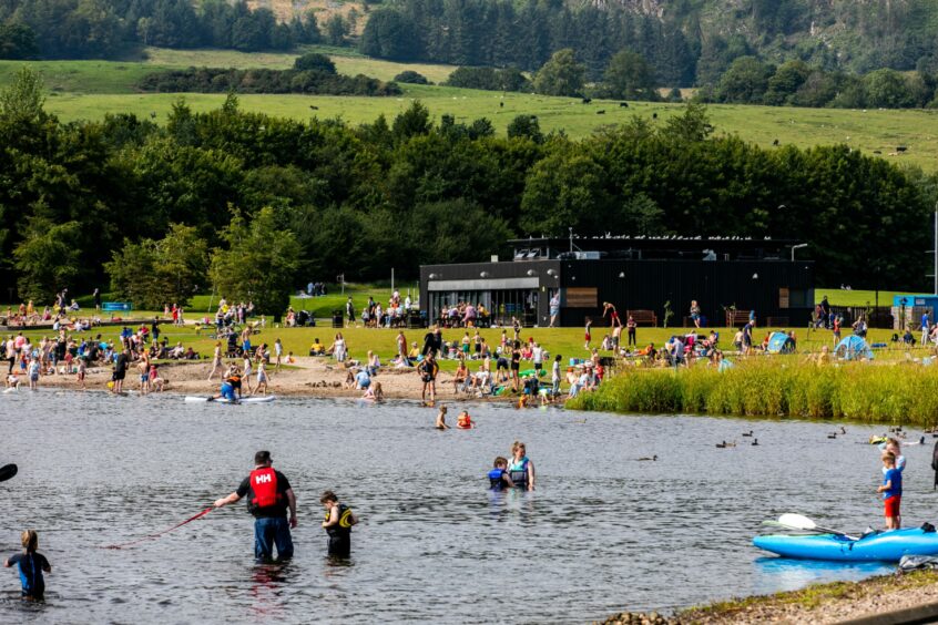 Image shows: Lochore Meadows on a busy sunny day. There are people enjoying the water in the loch and playing on the shore and in the playground in the distance.