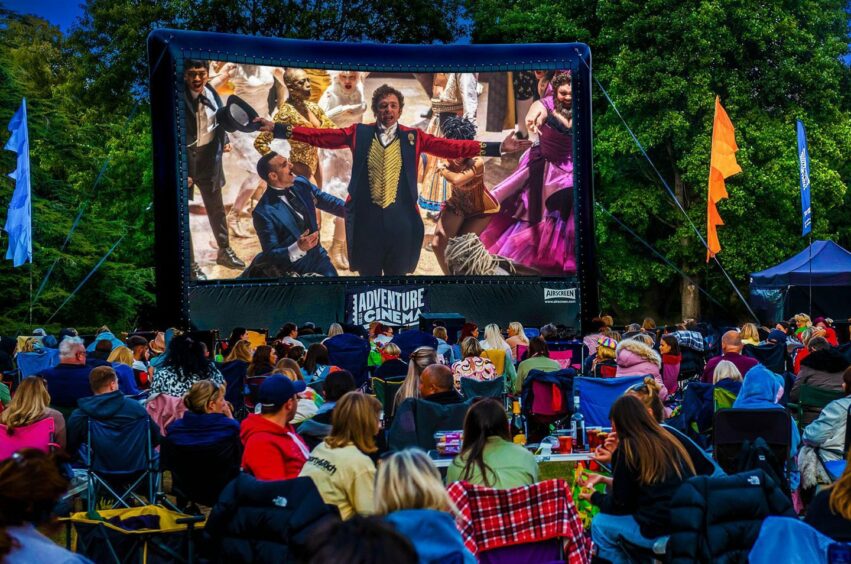 A large crowd enjoys an outdoor cinema showing the "Greatest Showman" film.