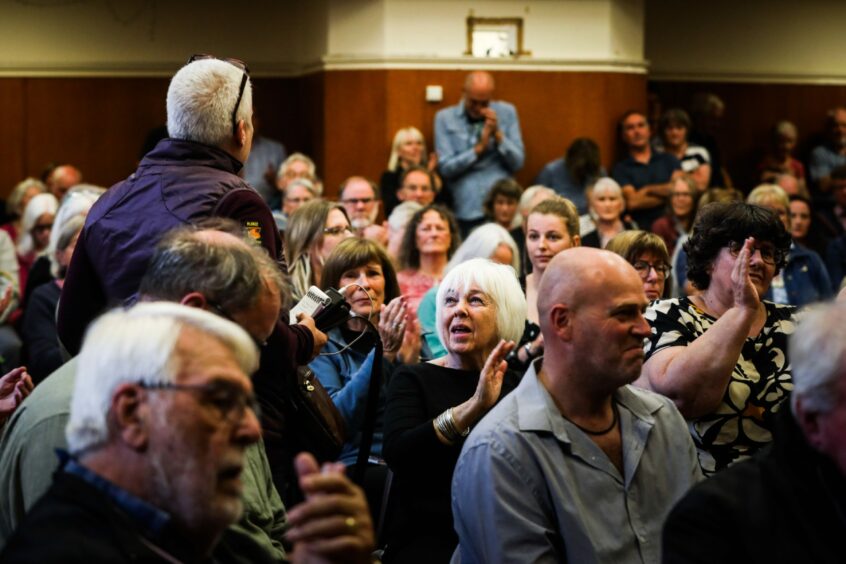 Faces in the crowd at the Aberfeldy public meeting on Discovery Land Company's plans for the Taymouth Castle estate. A man is on his feet speaking and people around him are applauding.