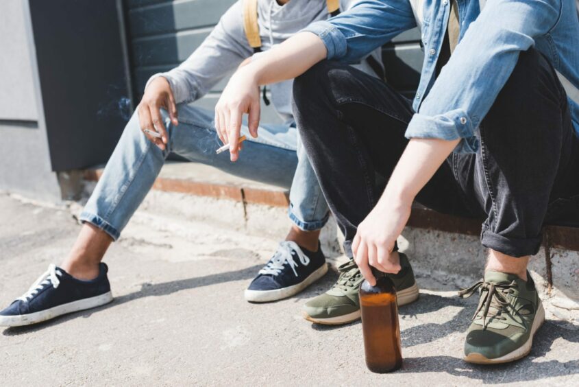 Two people, casually dressed and seated on a step. Both are smoking cigarettes, one is holding a beer bottle.