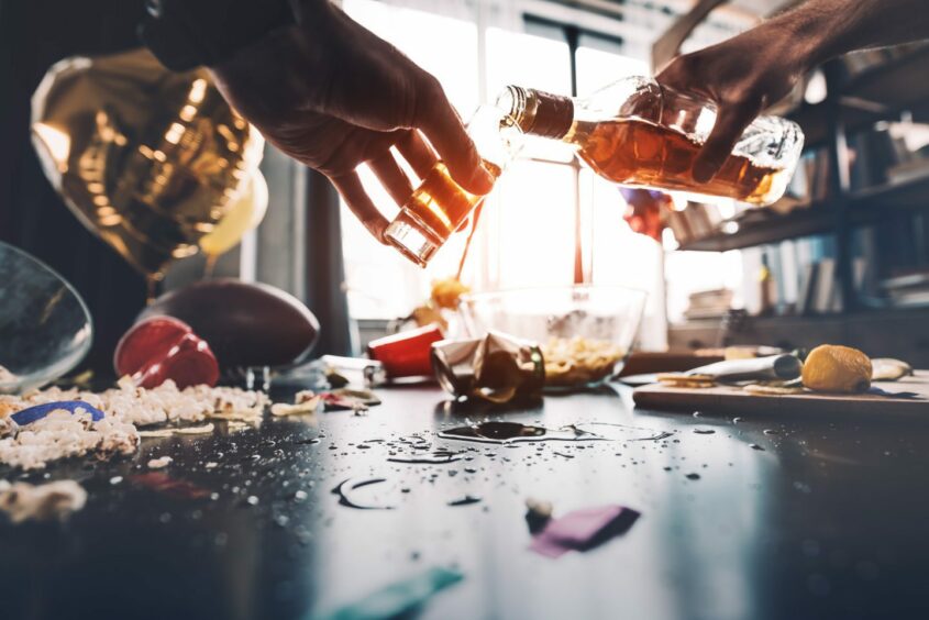 Two hands, one pouring alcohol into a glass held by the other, in front of the remains of a messy party.
