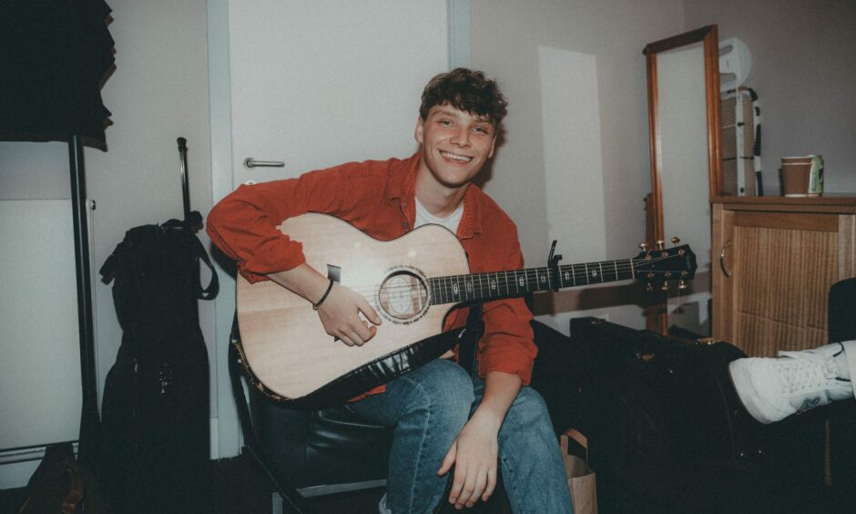 Ben Walker, wearing an orange button-down short over a white T-shirt and blue jeans, sits with his acoustic guitar in what appears to be a bedroom. 