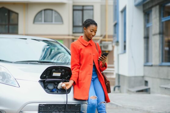 woman uses her smartphone and leans against her electric car as it charges