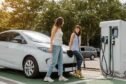 Two women talking to each other while their electric car charges at a charging station in a car park