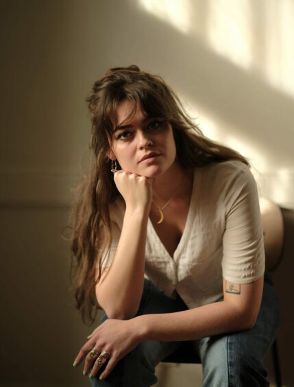 Poet Iona Lee is leaning forward on a chair with her chin resting on one hand. Her long brown hair is half tied up and she is wearing a white V-Necked top and gold necklace.