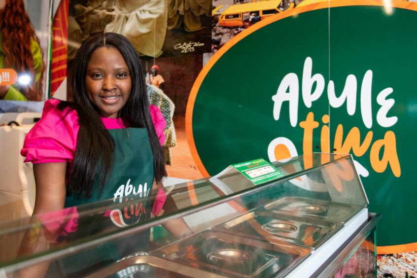 A woman wearing a pink tshirt and AbuleTiwa apron standing behind a takeaway counter.