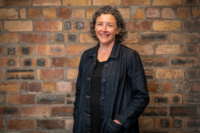 Image shows Helen O'Brien, Creative Director at Creative Catalyst in the studios. Helen is wearing trousers and a long jacket, has her hands in her pocket and is similing.