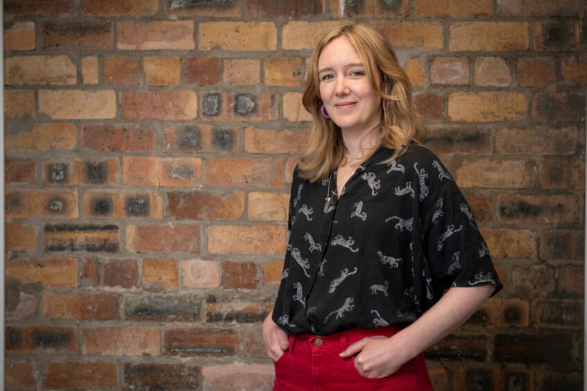 Image shows Anna Kelso, artist and creative facilitator at Creative Catalyst. Anna is standing in front of a red brick wall with her hands in her pockets. She is wearing red trousers and and a black patterned shirt.