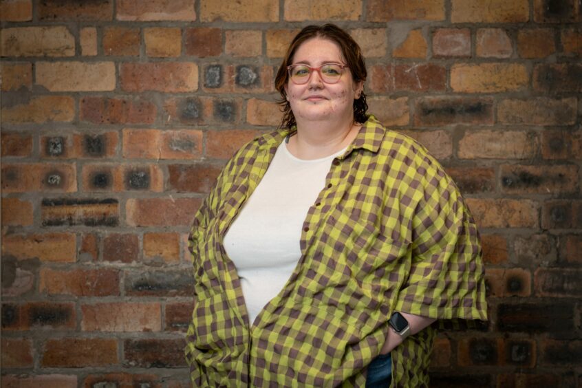 Image shows Caitlyn Hanna of Creative Catalyst. They are standing in front of a red brick wall and wearing a white t-shirt and black and yellow checked shirt.