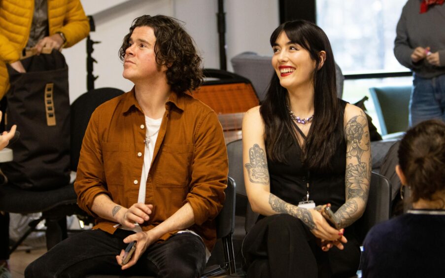 Kyle Falconer wears a tan button-down shirt, seated next to partner Laura Wilde, who is wearing a black outfit with silver chain and red lipstick.