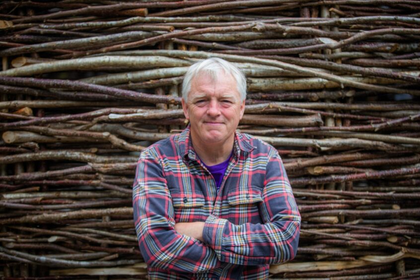 Scottish Crannog Centre director Mike Benson, arms folded in front of building woven from hazel branches.