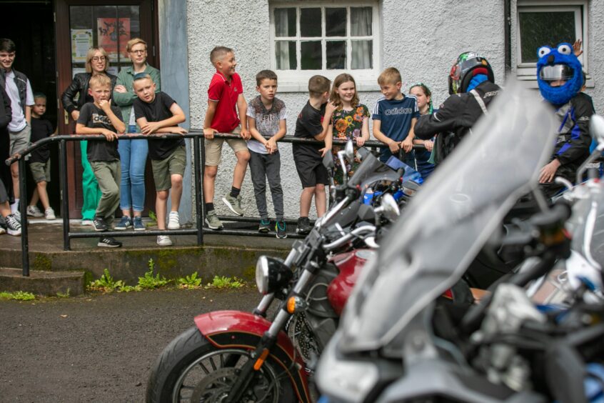 A group of children hanging over the railings outside Luncarty hall, as a group of motorbikes line up outside.