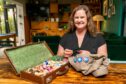 Image shows feature writer Nora McElhone sitting at a table with her Everyday Heirloom sewing case. The case is open showing colourful threads and she is sewing a Scout badge on an Explorers shirt.