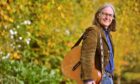 Dougie MacLean with guitar over his shoulder surrounded by autumn leaves.