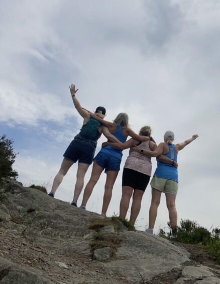 Four women lined up with backs to camera and hands in the air on a mountain top.