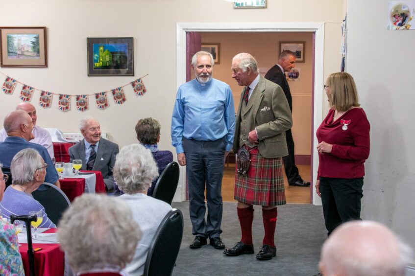 King Charles in kilt speaking to older people seated at a table in Kinross Day Centre with Union Jack bunting behind.