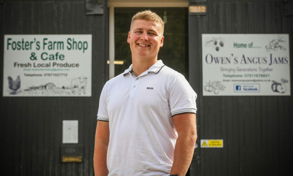 A 22-year-old man in a white tshirt in front of two signs saying Foster's Farm Shop and Cafe and Owen's Angus Jams.