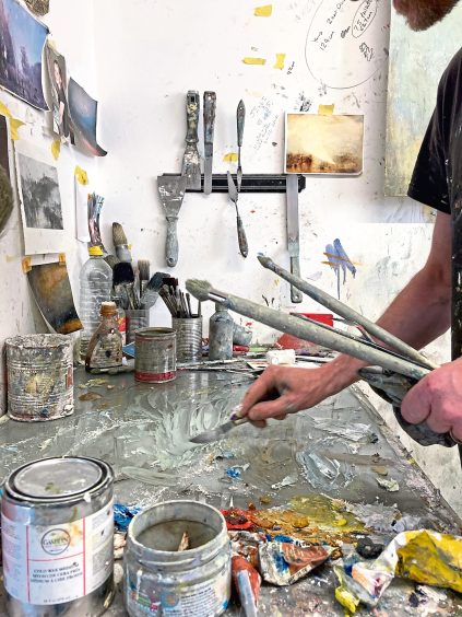 Image shows a close up of Robert Macmillan working on his new exhibition Light Half Hidden. Robert is holding brushes and mixing oil paint. Image: Robert Macmillan.