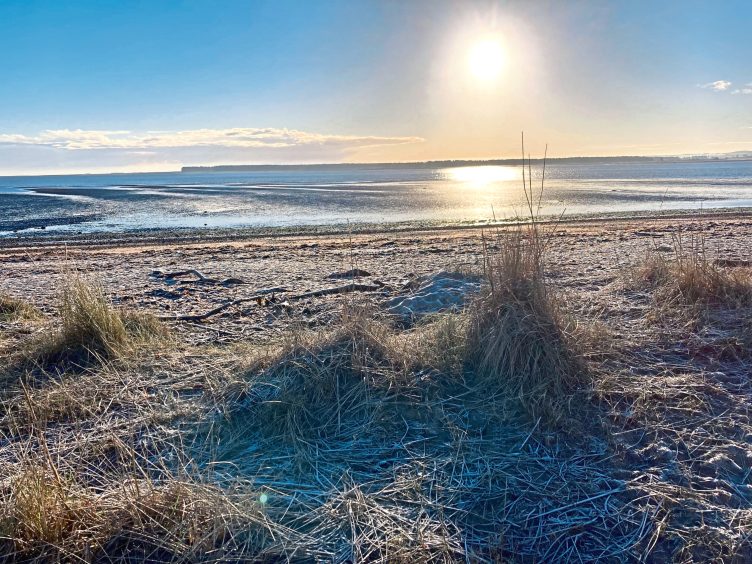 Image shows a photograph taken by artist Robert Macmillan while out walking on the coast. The sun is low in the sky and is reflected in the sea, with long shadows on the beach in the foreground.