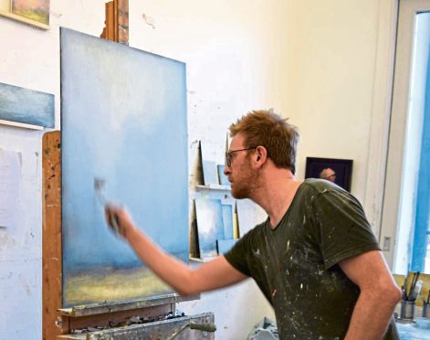 Image shows artist Robert Macmillan at work in his studio. He is working on a canvas on an easel and has paint on his clothes.