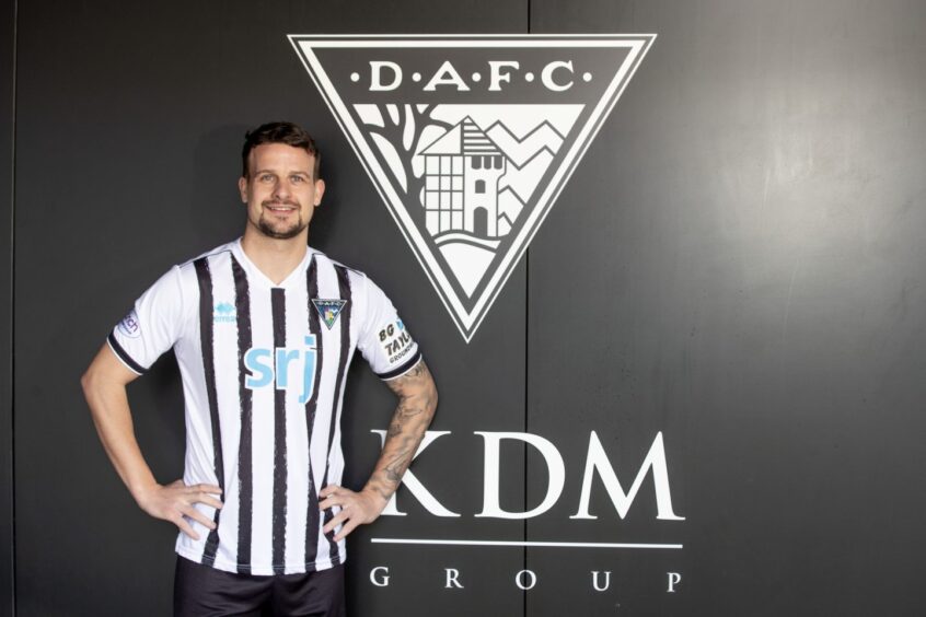 Kyle Benedictus stands beside a wall emblazoned with the Dunfermline Athletic F.C. badge.