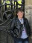 Image shows author Danielle Devlin who wrote Burnt Offerings standing in front of a black gate. Danielle has short dark hair and is wearing glasses. She is standing with her hands in her pockets and is wearing a t-shirt and black leather jacket.