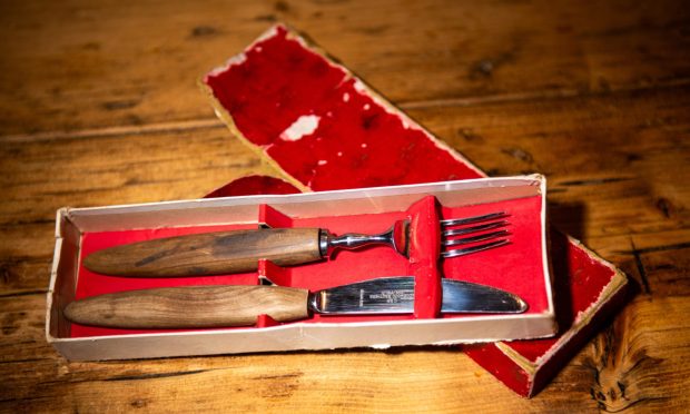 Image shows Catriona Steven's Everyday Heirloom, a steak knife and fork set. The knife and fork have stainless steel blade and prongs and wooden handles and are in a well-worn red presentation box.
