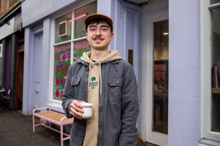 Barista Lewis standing outside the lilac shop front of EH9 Espresso with a coffee cup in hand.