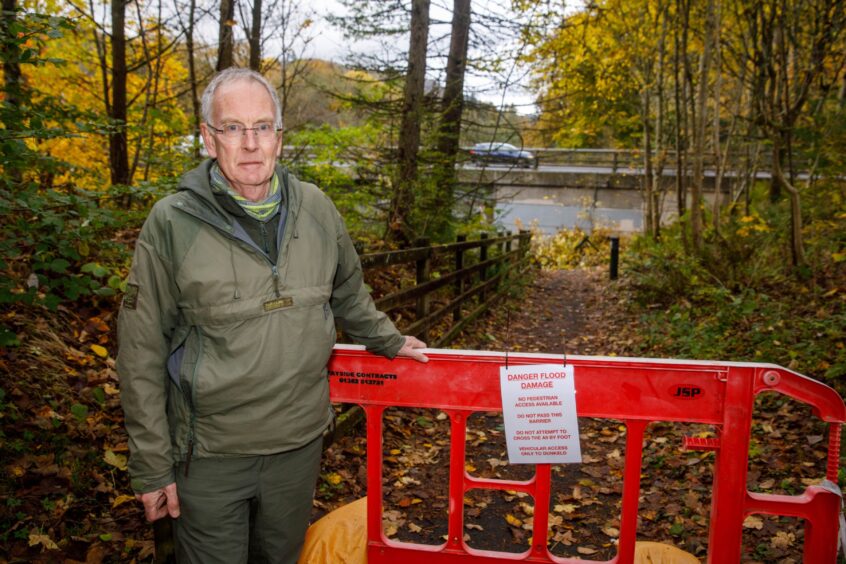 David Bee standing next to a barrier with a notice saying 'Danger, flood damage', beside the path leading the damaged underpass under the A9 at Dunkeld
