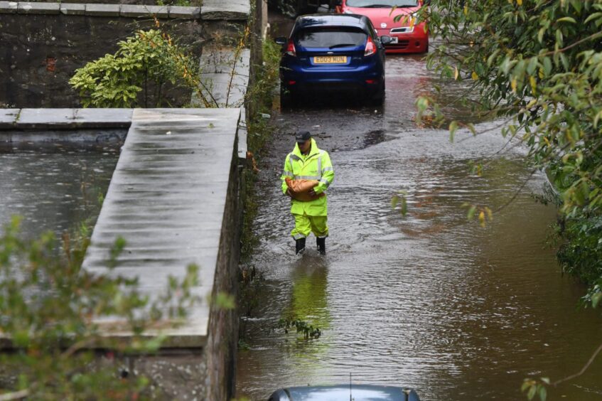 Man in high vis gear walking through puddles next to parked cars carrying a sandbag