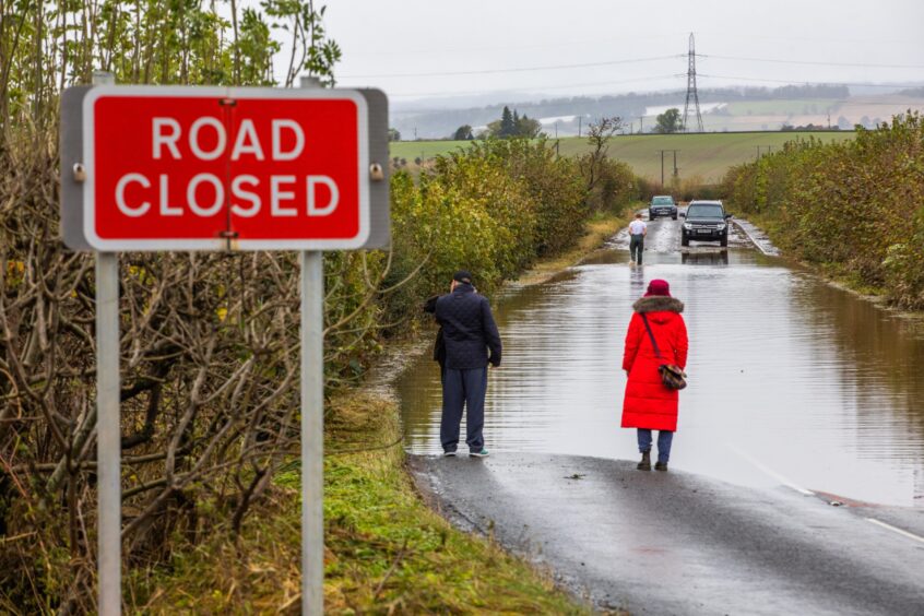 Two people with backs to camera watch a third person walking through flooded road to a vehicle on the far side. A 'road closed' sign is in the foreground.