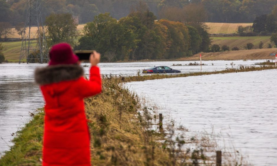 Person in red coat and hat taking photo of car submerged in flooded river Isla.