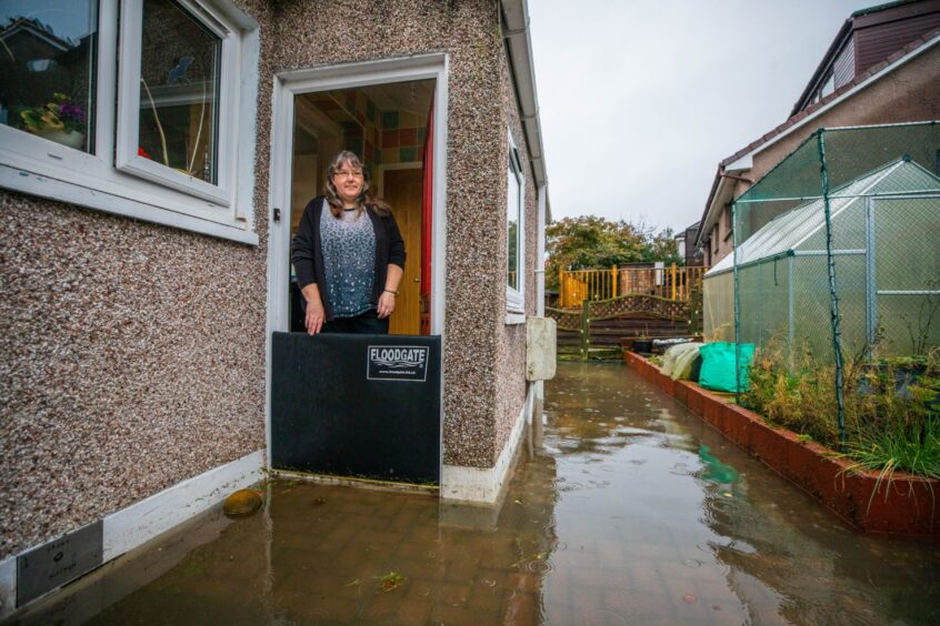 Janie Haig at the door to her home, with a flood gate across the threshhold and several inches of flood water outside.