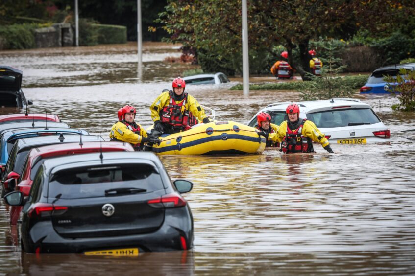 Four people in helmets and lifejackets steering inflatable boat past cares submerged in chest-deep floodwater in Invergwrie.