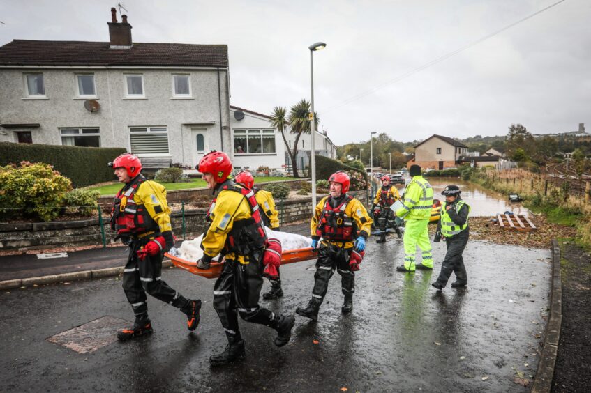 Rescuers carrying stretcher followed by a police officer walking away from flooded Invergowrie street