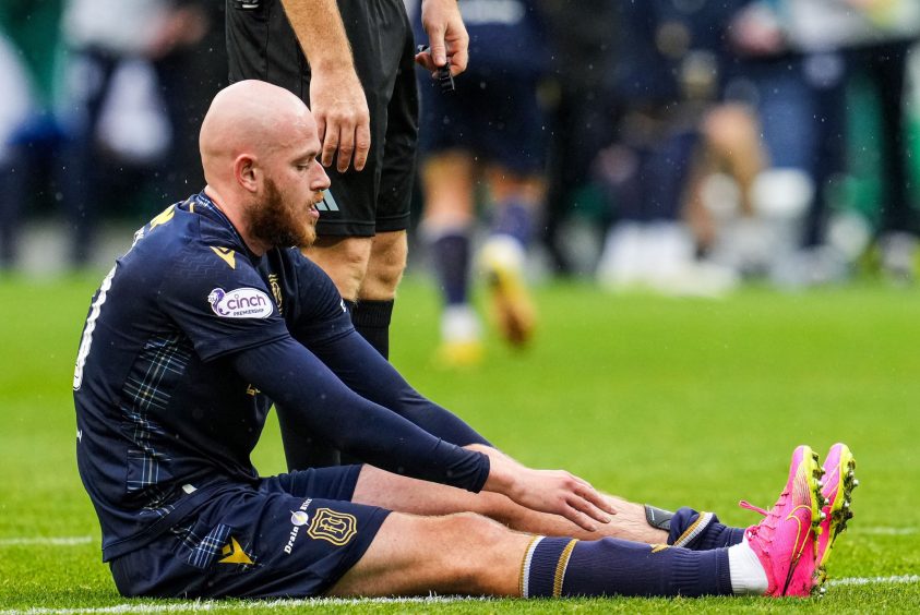 Dundee FC striker Zak Rudden sitting on the pitch, reaching down to touch his leg